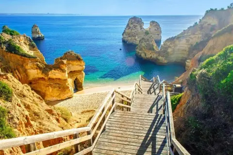 Schody prowadzą na piaszczystą plażę z turkusową wodą, idealne miejsce na portugalskie wakacje. Klify i skały tworzą malowniczy krajobraz.