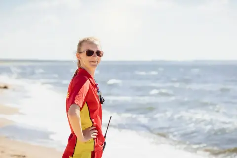 Livräddare i rött och gult står på stranden, redo för sol och bad. Vågorna skvalpar vid hennes fötter.