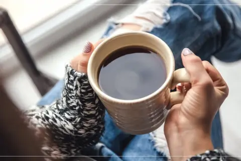 En person med slitna jeans och en stickad tröja håller en mugg med kaffe på morgonen.
