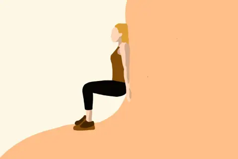 Kobieta w brązowym topie i czarnych legginsach wykonuje ćwiczenie wall sit, opierając plecy o ścianę.