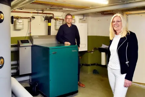 Ein Mann und eine Frau stehen neben einem grünen Nano BHKW-Gerät in einem Heizungsraum.