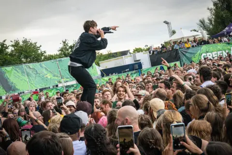 Sänger auf dem Dach eines Containers beim Green Juice Festival, der ins jubelnde Publikum zeigt. Viele Handys halten die Momente fest.
