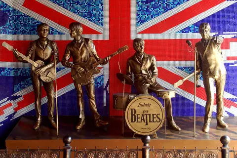 Pomnik The Beatles na tle flagi Wielkiej Brytanii. Czterech muzyków w pozłacanych strojach, z gitarami i perkusją, upamiętnia legendę muzyki.