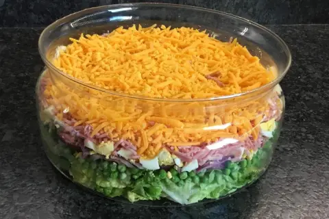 Une magnifique salade 7 étages dans un saladier en verre, avec des couches de laitue, petits pois, oignons rouges, jambon, œufs durs et fromage râpé.