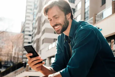 Uśmiechnięty mężczyzna z zarostem trzyma w dłoni czarny telefon, przeglądając coś z zaciekawieniem. W tle nowoczesne budynki.