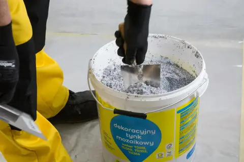 Ręka w czarnej rękawicy miesza łopatką dekoracyjny tynk mozaikowy weber w wiadrze.