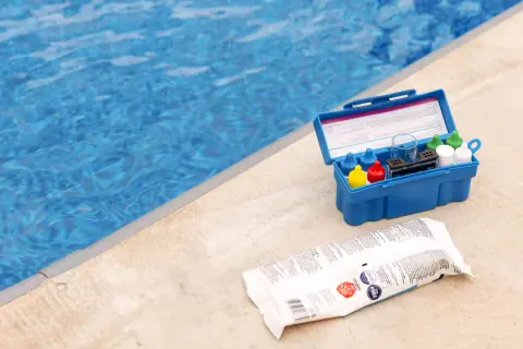Pool test kit and shock treatment next to a blue pool. Learn how to shock a pool for clear, clean water.