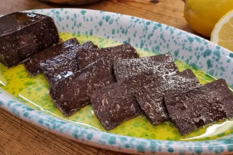Fette di salame di tonno scuro, lucido e saporito, servite in una ciotola con olio verde e limone.