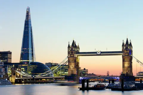 Widok na Tower Bridge i The Shard w Londynie o zmierzchu, z rzeką Tamizą i łodziami w tle.
