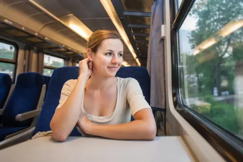 Uśmiechnięta kobieta podróżuje pociągiem za granicę, patrząc przez okno na mijane krajobrazy.