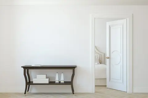 Un corridoio con un tavolino scuro e una porta aperta che rivela una stanza da letto. La porta interna è dipinta di bianco, con un design classico.