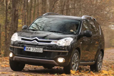 Czarny Citroen C-Crosser jedzie po leśnej drodze pokrytej jesiennymi liśćmi.