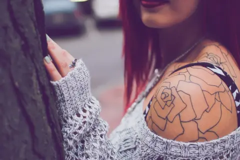 Dezent blühendes Tattoo Schulter Frau, eine zarte Rose ziert ihre Haut, umrahmt von einem Strickpullover.