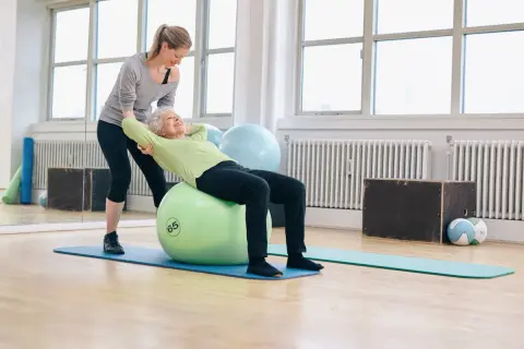 Une jeune femme aide une senior à faire des exercices sur un ballon de gym. Le **gym dos senior** est une activité bénéfique.