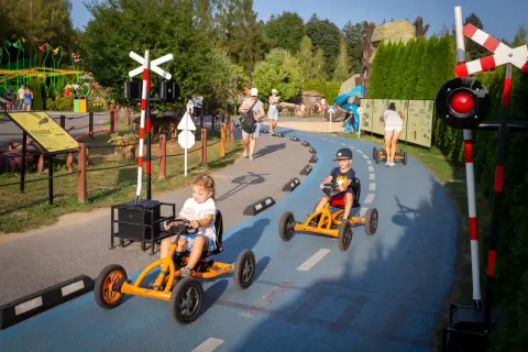 Dzieci ścigają się gokartami na torze w parku rozrywki. W tle widać inne atrakcje i spacerujących ludzi.