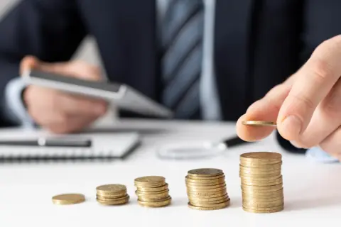 Dłoń układa monety, budując stosy. W tle mężczyzna z kalkulatorem, sugerującym planowanie finansowe i poszukiwanie optymalnych rozwiązań, być może związanych z **rajami podatkowymi**.