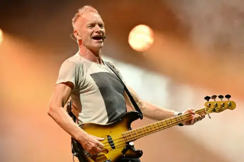 Sting, lider zespołu The Police, gra na gitarze basowej na scenie, oświetlony ciepłym światłem.