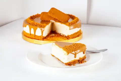 Ein Stück cremiger Lotus Cheesecake mit Karamell und Keksbröseln auf einem Teller, daneben der ganze Kuchen.