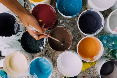 Ręka z pędzlem miesza farby w wiaderkach. Widać różne kolory, co może sugerować wady farby ceramicznej, np. nierównomierne krycie.