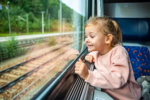Uśmiechnięta dziewczynka patrzy przez okno pociągu. Zastanawia się, od ilu lat można jeździć samemu pociągiem, podziwiając mijane krajobrazy.