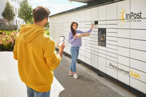 Mężczyzna z telefonem w ręku obserwuje kobietę, która wkłada paczkę do Paczkomatu InPost. Czy paczkomat InPost drukuje etykiety? Nie, ale ułatwia nadawanie.
