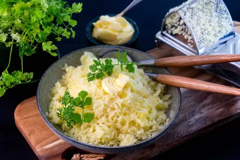 En skål med fluffig pressad potatis, toppad med en smörklutt och färsk persilja. Bredvid syns en smörskål och en rivare.