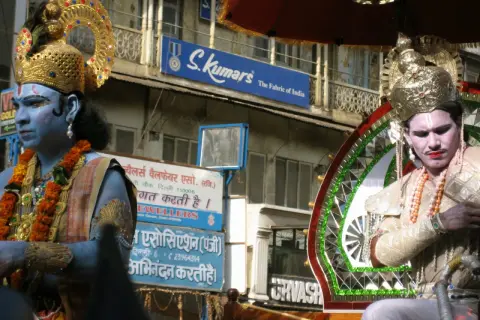 Två män i gyllene krön och smink, en med blå hud, deltar i en procession. Frågan "hur många gudar har hinduismen" kan väckas av deras utseende.