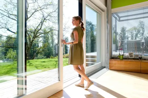 Kobieta w letniej sukience otwiera drzwi, wychodząc na taras. Duże okna wpuszczają mnóstwo światła do nowoczesnego wnętrza.