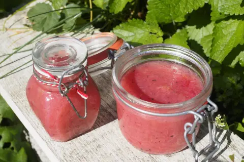 Två glasburkar med rabarbermarmelad med kardemumma står på en träbänk utomhus, omgivna av gröna blad.