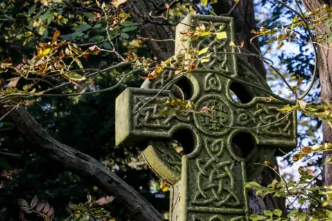 Kamienny krzyż celtycki z misternymi wzorami, otoczony jesiennymi liśćmi. Symbolizuje wieczność i połączenie życia.