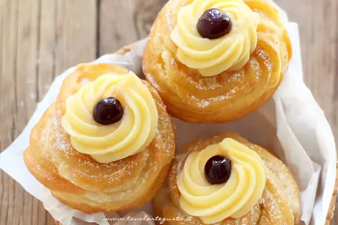 Tre zeppole fritte napoletane, dorate e spolverate di zucchero a velo, con crema pasticcera e una ciliegina.