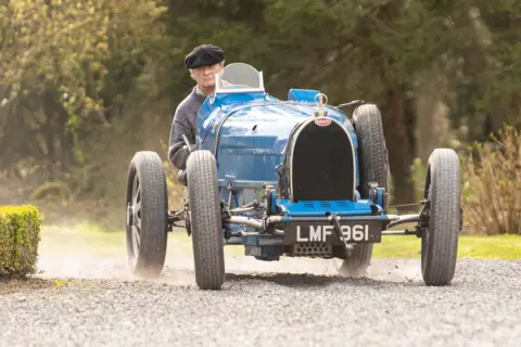 Ein Mann in einem schwarzen Hut fährt einen blauen Bugatti Type 35 über einen Schotterweg.