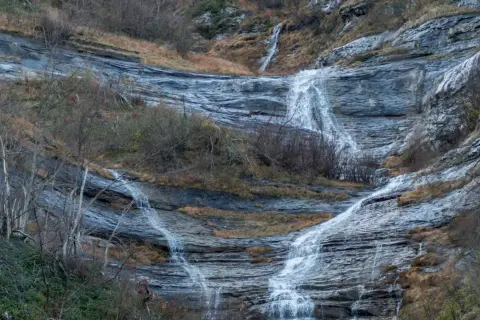 Le cascate della Volpara scendono su rocce stratificate, con vegetazione autunnale e alberi spogli che incorniciano il paesaggio.