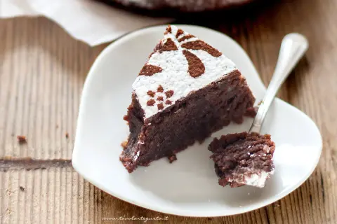 Una fetta di torta caprese ricetta facile, cosparsa di zucchero a velo e cacao, su un piatto bianco a forma di cuore. Un cucchiaino ha prelevato un pezzetto.
