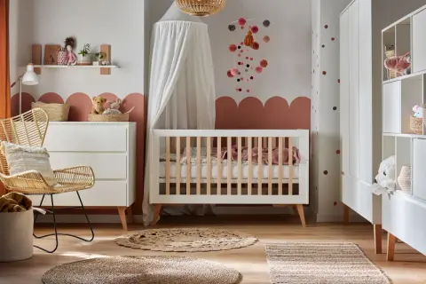 Przytulny pokój niemowlaka z białym łóżeczkiem, wiklinowym fotelem i komodą. Dekoracje w pastelowych kolorach tworzą ciepłą atmosferę.