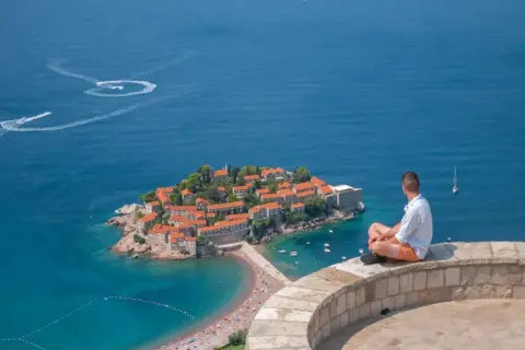 Mężczyzna siedzi na murze, podziwiając wyspę Sveti Stefan. Czy Czarnogóra jest bezpieczna? Tak, to spokojne miejsce na wakacje.