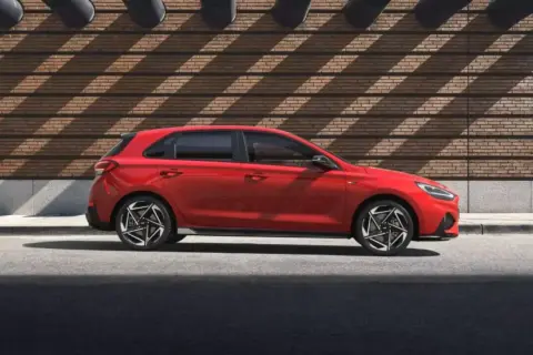 Czerwony Hyundai i30 Hatchback zaparkowany na tle ceglanej ściany z geometrycznymi cieniami.