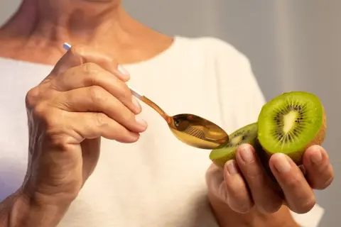 Dłonie trzymają połówkę kiwi i złotą łyżeczkę. Owoc jest bogaty w witaminy, np. C i K.