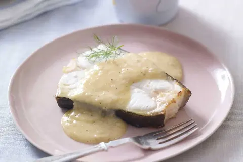 En bit fisk täckt med krämig beurre blanc-sås, garnerad med dill, serveras på en rosa tallrik med en gaffel.