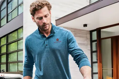 Mann trägt ein blaues Henley Shirt mit Knopfleiste und rotem Logo. Was ist ein Henley Shirt? Ein lässiges Oberteil mit Kragen und Knopfleiste.