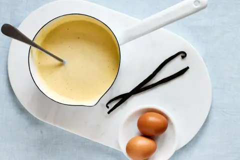 Zutaten für vanillesauce selber machen: ein Topf mit cremiger Sauce, zwei Eier und zwei Vanilleschoten.