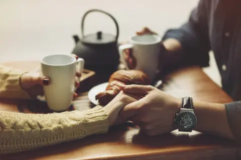 Dłonie trzymające kubki z kawą i croissant, symbolizujące czułe komplementy dla kobiety.