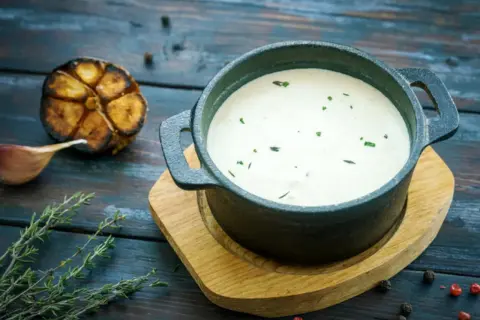 Cremoso alioli con leche, servido en cazuela de hierro fundido, acompañado de ajo asado y tomillo fresco.