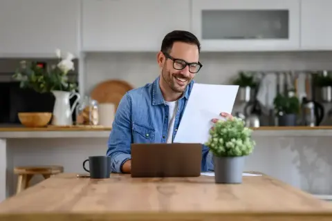 Uśmiechnięty mężczyzna w okularach przegląda dokumenty przy laptopie. W skład jego domowego biura wchodzi kubek i roślina.