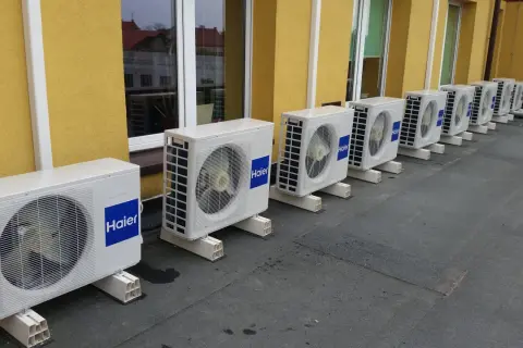 Rząd białych jednostek klimatyzacyjnych Haier na dachu, na tle żółtej ściany budynku.