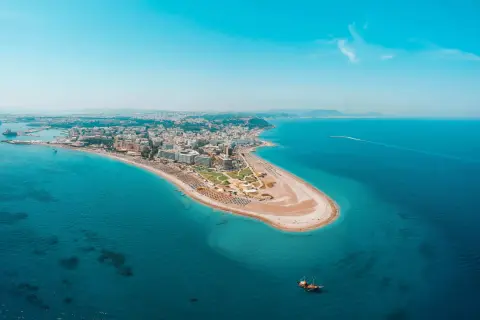 Luftfoto över Rhodos kustlinje med en sandstrand, hotell och turkost vatten. En segelbåt syns i vattnet, perfekt för Rhodos aktiviteter.