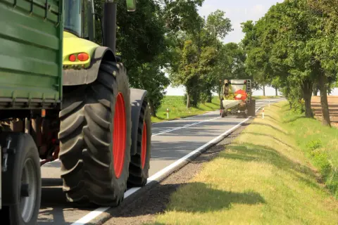 Ciągnik z przyczepą przygotowuje się do wyprzedzania pojazdu wolnobieżnego na wiejskiej drodze.