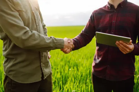 Dwóch mężczyzn w polu ściska sobie dłonie, jeden z nich trzyma tablet. Rozmowa o tym, jak sprawdzić, czy działka jest uzbrojona.