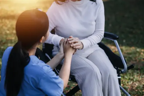 Un infermiere tiene le mani di una paziente su una sedia a rotelle, un gesto di conforto e cura. L'età non conta, l'assistenza è l'essenza.