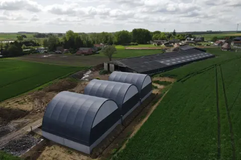 Tre tunnel stalla grigi, moderni, affiancati in un campo verde, con un grande edificio agricolo sullo sfondo.